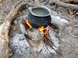Traditional village cooking setup and makeshift stove. Water being heated over open fire. Rustic surroundings, dry leaves, branches. Evokes nostalgia, connection to simpler village life. photo
