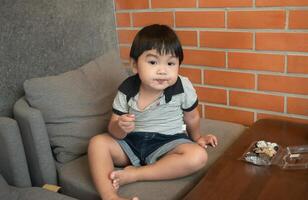 Asian cute little boy sitting on the sofa using a plasic spoon to eat mini donuts at home,enjoys eating looking at the camera and happiness photo