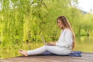 A woman is sitting on a dock with a laptop open in front of her. She is wearing white pants and a white sweater. The scene is peaceful and relaxing, with the water in the background. photo