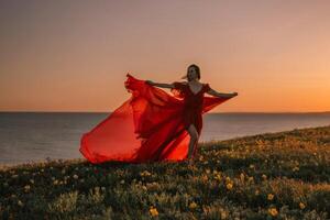 woman red dress is standing on a grassy hill overlooking the ocean. The sky is a beautiful mix of orange and pink hues, creating a serene and romantic atmosphere. photo