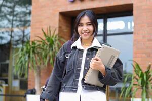 portrait of smiling asian woman holding laptop and carrying bag walking out from cafe photo