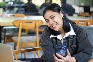 cheerful asian woman holding smartphone to surfing in social media while working remote from cafe using laptop looking at camera photo