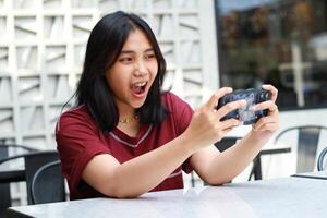 pretty asian woman excited playing game online using mobile phone sitting in outdoor cafe wearing red tshirt, shouting with open mouth photo