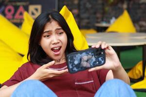 shocked asian woman defeated playing game using smartphone in outdoor cafe sitting in bean and covering her mouth photo