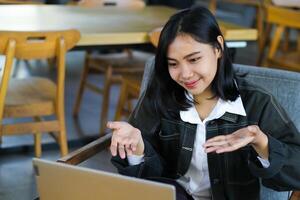 pretty asian woman looks happy doing virtual presentation with client in cafe using laptop, raising palms showing presenting gesture photo