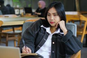 depressed asian woman writing in notebook using pen and daydreaming while work in indoor cafe using laptop photo