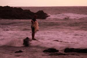a man standing on rocks in the ocean with a surfboard photo