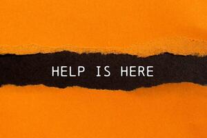 Help is here message written on ripped torn orange paper on black background. Conceptual help is here symbol. Copy space. photo