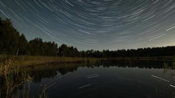Witness the mesmerizing trails of stars glowing in the night sky above a calm lake surrounded by trees. A breathtaking display of nature's beauty unfolds as time passes. video