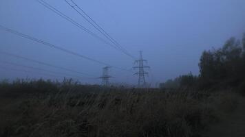 A fog-covered landscape reveals towering power lines stretching across a desolate field. The dense mist creates a mysterious ambiance as day transitions to night. video