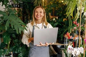 Smiling florist working on laptop and taking online orders in store and looking at camera photo