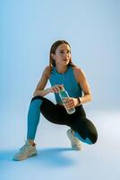 Active woman drinking water from bottle while workout and doing sport exercises on studio background photo