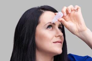 Woman applying eye drops against gray background for eye hydration and relief. photo