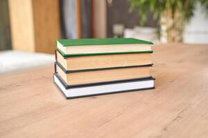 Stack of hardcover books on wooden table in cozy indoor setting with greenery and natural lighting. photo