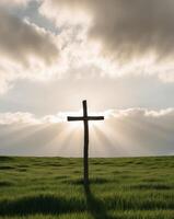 A tight shot of a cross standing alone in a sea of green grass Sun rays filtering through clouds behind. photo