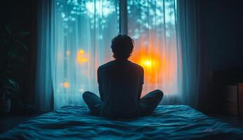 A man is sitting on a bed with his back to the camera. The room is dimly lit, and the man is looking out the window. Scene is contemplative and peaceful photo