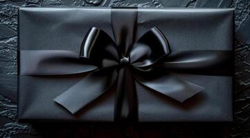A black box with a bow on top of it. The box is wrapped in black paper and has a black ribbon tied around it. The bow is tied in a bow shape and is positioned in the center of the box. photo