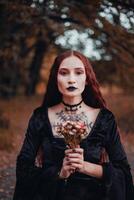 Halloween witch, woman in a black dress holds a colorful bouquet in a forest at dusk photo