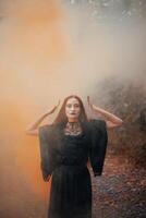 Goth woman, red-haired witch in autumn forest in fog in black dress, Halloween. Mystical atmosphere photo