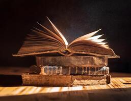 A stack of weathered books with an open one on top, illuminated by warm light, highlighting dust particles in the air. photo