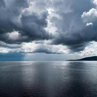 Dramatic dark storm clouds hover over calm ocean waters, creating a moody and serene seascape with distant land on the horizon. photo