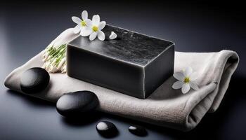 A black charcoal soap bar resting on a towel, accompanied by white flowers and spa stones, creating a tranquil spa atmosphere. photo