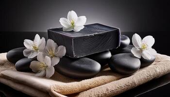 Charcoal soap bar displayed with white flowers and smooth black spa stones on a textured towel, creating a serene spa setting. photo