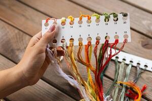 Hand holding a card with numbered slots for different colored embroidery threads. Card and thread are laid out on a wooden surface, suggesting a craft setting. Embroidery. photo