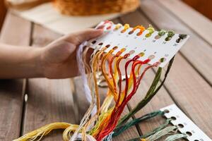 Hand holding a card with numbered slots for different colored embroidery threads. Card and thread are laid out on a wooden surface, suggesting a craft setting. Embroidery. photo
