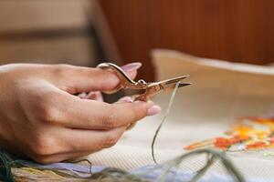 A hand holding embroidery scissors about to cut a thread. A hand with scissors cuts the thread. Close-up of cross stitch. Crafts and embroidery. photo