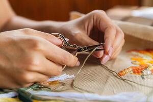 A hand holding embroidery scissors about to cut a thread. A hand with scissors cuts the thread. Close-up of cross stitch. Crafts and embroidery. photo
