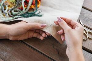 A woman doing cross stitching. Close-up of hands with a needle threading the thread through the fabric. Embroidery of a picture. In the background are colorful embroidery threads. photo