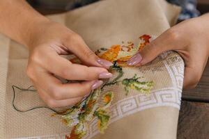 A woman doing cross stitching. Close-up of hands with a needle threading the thread through the fabric. Embroidery of a picture. In the background are colorful embroidery threads. photo