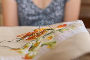 A woman doing cross stitching. Close-up of hands with a needle threading the thread through the fabric. Embroidery of a picture. In the background are colorful embroidery threads. photo