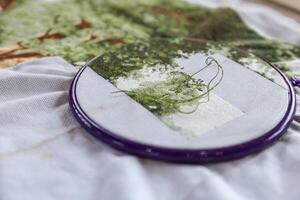 Close-up of a cross-stitch embroidery project in progress. The needle is threaded with green floss and is partially inserted in the fabric. The hoop is purple, and the fabric is white. photo