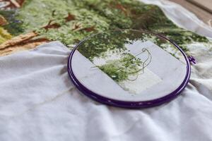 Close-up of a cross-stitch embroidery project in progress. The needle is threaded with green floss and is partially inserted in the fabric. The hoop is purple, and the fabric is white. photo