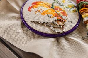 Cross-stitch, with an embroidery hoop containing a partially completed design of an orange and white flower. A pair of gold embroidery scissors rests on top of the hoop. photo