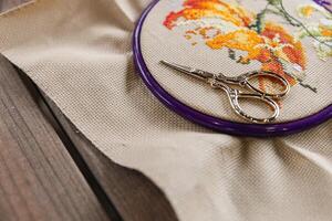 Cross-stitch, with an embroidery hoop containing a partially completed design of an orange and white flower. A pair of gold embroidery scissors rests on top of the hoop. photo