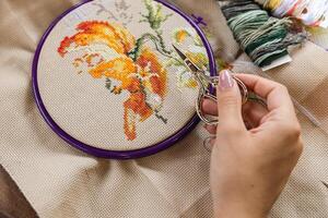 Person working on a cross-stitch. They are using a pair of small, gold-colored scissors to trim the threads. The embroidery hoop, cross-stitch design is partially shown, featuring a floral motif. photo