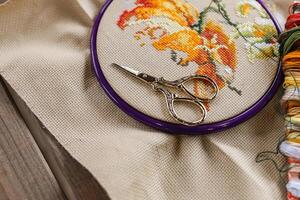 Cross-stitch, with an embroidery hoop containing a partially completed design of an orange and white flower. A pair of gold embroidery scissors rests on top of the hoop. photo