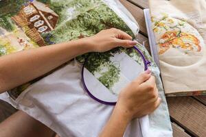 The image shows a person working on a cross-stitch embroidery project. They are using an embroidery hoop to hold the fabric taut and are carefully guiding the needle through the cloth. Hobbies. photo