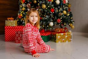blonde girl in red pajamas is sitting on the floor next to a beautifully decorated Christmas tree photo
