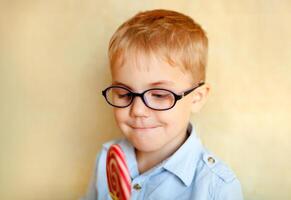 A small boy with strabismus in glasses is happily eating a big red lollipop photo