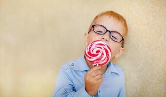 A young boy with glasses and squinting eyes is delightfully munching on a big red lollipop. photo