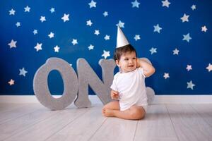 Baby boy of one year old wearing a white body and a festive paper hat sits on the floor on a blue background with stars and big silver letters photo