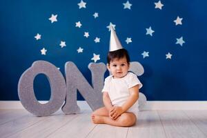 Baby boy of one year old sits on the floor on a blue background with stars and big silver letters one photo