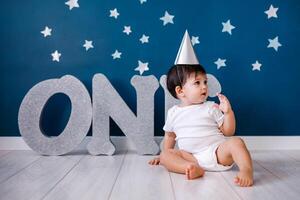Baby boy of one year old wearing a white body and a festive paper hat sits on the floor on a blue background photo