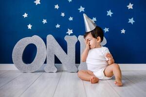 Baby boy of one year old wearing a white body and a festive paper hat sits on the floor photo