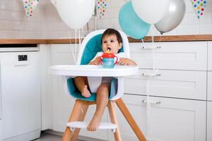 Little baby boy sitting in high chair at home on white kitchen and drinking water from sippy cup on background with balloons. photo