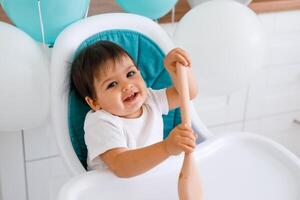 Little baby boy sitting in blue high chair at home on white kitchen and playing with wooden big spoon on background with balloons. photo
